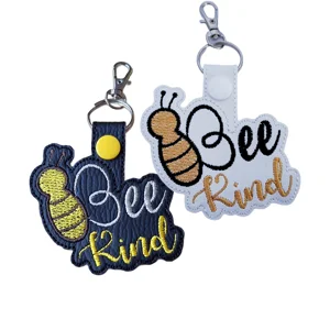 Bee Kind embroidered faux leather keyrings in black and white, with bee design, stitched lettering and metal clips.