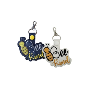 Two Bee Kind embroidered keyrings shown side by side, one in black and one in white faux leather, each featuring a bee design with the words ‘Bee Kind’ stitched in contrasting thread and finished with a metal clasp and keyring.
