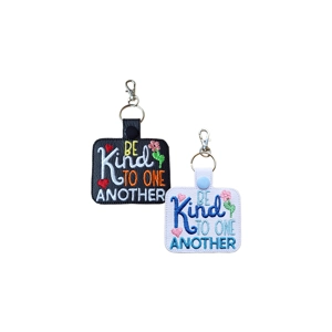 Two faux leather ‘Be Kind to One Another’ keyrings shown side by side. One is black and the other white, each featuring embroidered text with heart and flower details, a rectangular shape, and a metal keyring with lobster clasp.