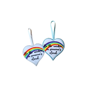 Two heart-shaped hanging decorations with embroidered rainbows and the message ‘Be the rainbow in someone’s cloud’. Each heart has a stitched border and a ribbon loop for hanging.