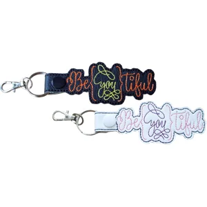 Be You Tiful embroidered faux leather keyrings in black and white, with colourful stitched lettering and metal clips.