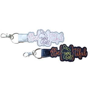 Two BeYOUtiful embroidered keyrings shown side by side, one in white faux leather and one in black faux leather, each featuring decorative stitched lettering, finished with a metal lobster clasp and keyring.