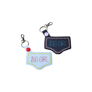 Two faux leather keyrings shaped like a pair of knickers. One is white with yellow stitching and red 'BIG GIRL' text, finished with a red snap. The other is black with pink stitching and blue 'BIG GIRL' text, finished with a lilac snap. Both have metal keychain hardware attached.