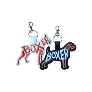 Two Boxer dog silhouette keyrings shown side by side, made from faux leather with embroidered ‘Boxer’ lettering. One design is white with brown embroidery and the other is black with white and brown embroidery, each finished with a metal clasp and keyring.