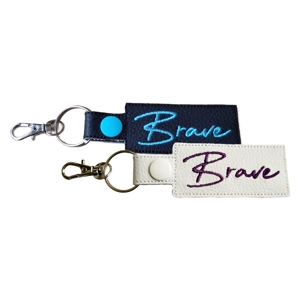 Two faux leather affirmation keyrings embroidered with the word ‘Brave’, shown in black and white colour options with metal keyring hardware.