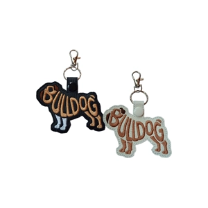 Two embroidered bulldog-shaped keyrings made from faux leather. One is black with tan word-art stitching that spells 'Bulldog', and the other is white with the same design. Both keyrings have snap fasteners and metal swivel clasps attached at the top.