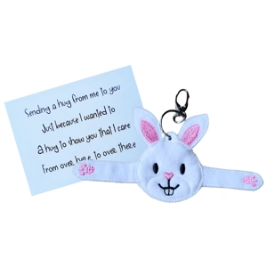 A soft felt bunny pocket hug keyring with outstretched arms, machine embroidered details, and a silver keyring clip, shown alongside a printed keepsake card.