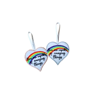 Two heart-shaped decorations with hanging white ribbons, each embroidered with a rainbow and the message ‘You are capable of amazing things’. One has a soft pink border and the other has a navy blue border.