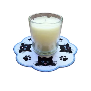 Embroidered cat and paw print candle mat with stitched cat faces, paw prints, stippled pattern and scalloped edge, shown with a candle in the centre.