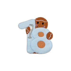 A felt gingerbread character holding a light blue number 18, with stitched details, a smiling face, and a small champagne glass.