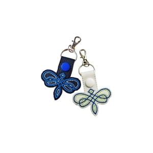 Two faux leather dragonfly-shaped keyrings featuring embroidered Celtic-inspired designs. One keyring is black with blue-toned stitching, and the other is white with green and blue stitching, each finished with a metal keyring and lobster clasp.