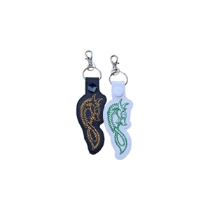 Two faux leather dragon-shaped keyrings featuring embroidered Celtic-inspired guardian dragon designs. One keyring is black with gold stitching, and the other is white with green stitching, each finished with a metal keyring and lobster clasp.