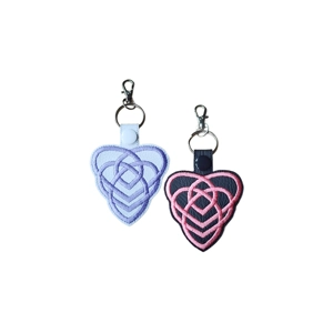 Two Celtic Heart of Motherhood keyrings made from faux leather, one white with lilac embroidery and one black with pink embroidery. Each keyring features an interwoven Celtic heart knot and a lobster clasp at the top.