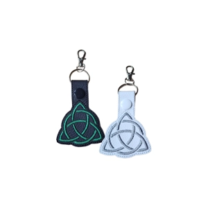 Two Celtic Triquetra Keyrings on a white background. One keyring is black with green embroidery, the other is white with grey embroidery. Both feature a triquetra knot design and a metal keyring with a lobster clasp.