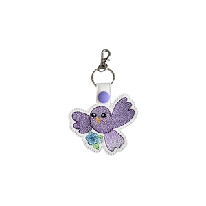 A kawaii-style bird keyring made from faux leather, featuring a purple embroidered bird with outstretched wings, bright eyes, and a small blue flower detail, finished with a metal keyring and lobster clasp.