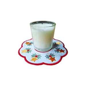 A white candle in a glass holder placed on a scalloped white embroidered Christmas mat featuring golden star ornaments, green holly with red berries, and a red stitched border.