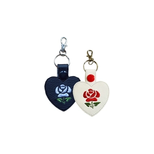 Two heart-shaped faux leather keyrings, one black and one white, each with an embroidered rose and green leaves. The black keyring has a pale blue rose with a black snap and silver clasp, while the white keyring has a red rose with a red snap and bronze clasp.
