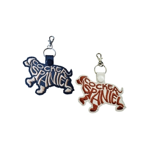 Two embroidered Cocker Spaniel silhouette keyrings shown on a white background. One keyring is black with cream stitching, the other is white with brown stitching. The word Cocker Spaniel is stitched repeatedly to form the shape of the dog. Each keyring has a metal clasp and ring attached at the top.