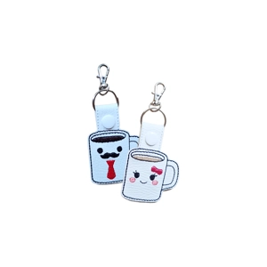 Two faux leather coffee mug keyrings with smiling faces. One mug has a red tie and moustache, the other has a pink bow and rosy cheeks, each attached to a silver keyring clip.