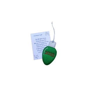 A green felt ornament shaped like a classic Christmas light bulb embroidered with “2023” in red, attached to a silver top and hanging loop. Behind it is a printed keepsake card titled “A Colleague’s Light,” featuring a short festive poem expressing appreciation for a colleague.
