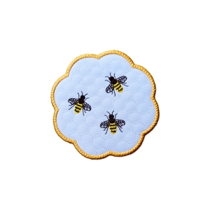 Scalloped embroidered candle coaster featuring three honeybee motifs stitched on a softly textured background, finished with a contrasting embroidered border.