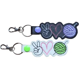 Two Crochet Peace Love faux leather keyrings in black and white, featuring embroidered peace sign, heart and yarn ball designs.