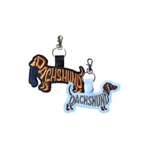 Two embroidered Dachshund keyrings shown side by side on a white background. One is black with orange stitching and a small blue lead detail, the other is light grey with brown stitching. Both are dog-shaped word-art designs with metal lobster clasps and keyrings attached.