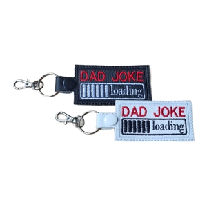 Two faux leather dad joke keyrings, one black and one white, featuring an embroidered loading bar design with the words Dad Joke Loading, finished with a metal clasp and keyring.