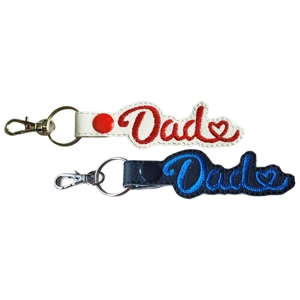 Two faux leather Dad keyrings, one in white with red embroidery and one in black with blue embroidery, each featuring the word 'Dad' stitched in cursive with a small heart detail and finished with a metal ring and lobster clasp.
