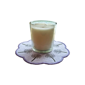 A white candle in a glass holder stands on a scalloped white embroidered mat decorated with delicate dandelion seed heads and a pale lilac stitched border.