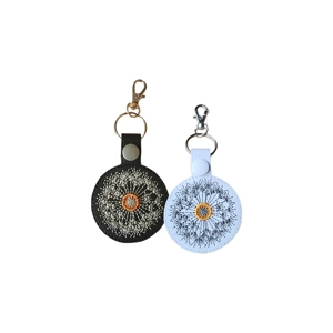 Two round faux leather keyrings with embroidered dandelion clock designs. One is black with cream stitching and a gold centre, and the other is white with dark grey stitching and a yellow centre. Each keyring has a snap tab and metal clip.