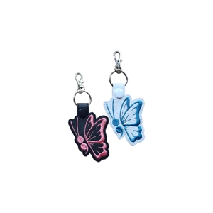 Two faux leather butterfly keyrings with simple stitched wing designs. One keyring is black with pink embroidery and a black snap, and the other is white with blue embroidery and a white snap. Both have silver keyrings and lobster clasps attached at the top.