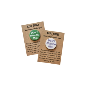 Two round magnetic needle minders on kraft instruction cards, one with a green background and white text and one with a white background and purple text, both featuring the words “Don’t Needle Me!”.
