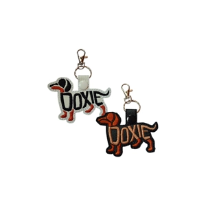 Doxie embroidered keyring shown in black and white faux leather options, featuring a dachshund silhouette with stitched lettering and a metal lobster clasp.