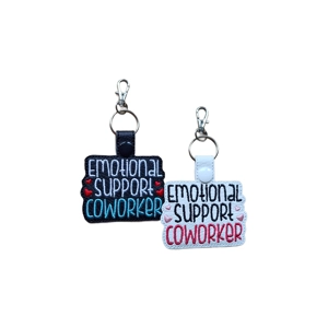 Two faux-leather keyrings with embroidered text reading 'Emotional Support Coworker.' One keyring has a black base with white, teal, and red stitching, and the other has a white base with black, pink, and red stitching. Both feature small embroidered hearts and have a snap tab with a silver keyring and lobster clasp at the top.
