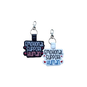 Two faux-leather keyrings embroidered with the phrase 'Emotional Support Human'. One keyring is black with white, pink, and blue stitching and small red hearts; the other is white with black, blue, and red stitching and small pink hearts. Both have a snap tab and a silver keyring with a lobster clasp.
