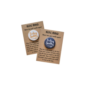 Two round “Feeling Stabby” needle minders displayed on brown kraft backing cards. One has an orange design on a white background, the other white text on a navy background, both with a small needle graphic and magnetic backing.