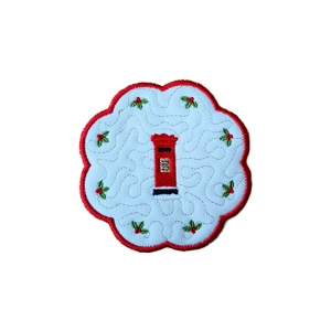 Round scalloped coaster mat with an embroidered red post box in the centre and green holly sprigs with red berries around it on a white background, featuring stitched details and a red embroidered border.