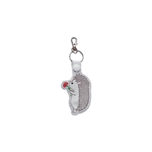 A faux leather hedgehog keyring with detailed machine embroidery, showing a small hedgehog holding and sniffing a flower, finished with a metal keyring and clip.