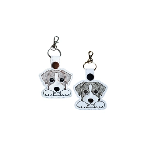 Two kawaii-style long-haired Jack Russell dog keyrings made from faux leather, featuring a fluffy puppy design with paws-up pose and machine embroidery, shown in black and white and brown and white versions with metal keyrings and lobster clasps.