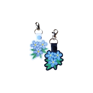 Two faux leather forget-me-not flower keyrings with detailed machine embroidery, shown in black and white colour options with metal keyring and lobster clasp attachments.