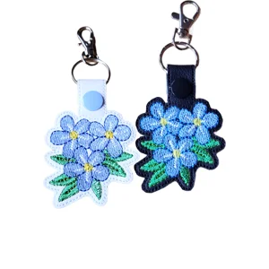 White and black faux leather forget me not flower keyrings with blue embroidered flowers, green leaves and silver metal clasp hardware.