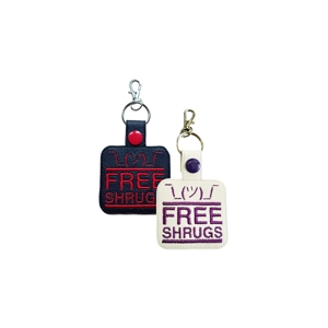 Two faux leather Free Shrugs keyrings with machine embroidered text, shown in black with red stitching and white with purple stitching, each finished with a metal keyring and lobster clasp.
