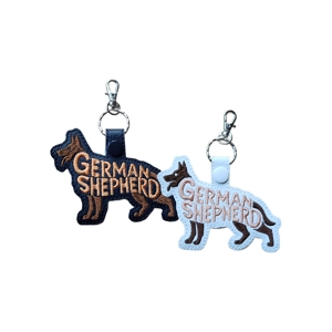 Two German Shepherd–shaped faux leather keyrings with embroidered word art. One is white with black stitching and a white snap tab, the other is black with tan stitching and a black snap tab. Both have silver keyrings and lobster clasps attached at the top.