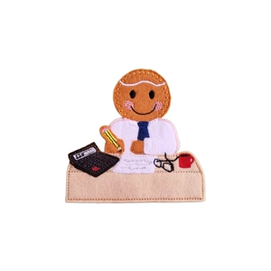 A felt gingerbread character dressed as an accountant, sitting at a desk with a pencil, calculator, papers, glasses, and a red mug. The design is machine embroidered with fine stitched details in soft felt fabric.