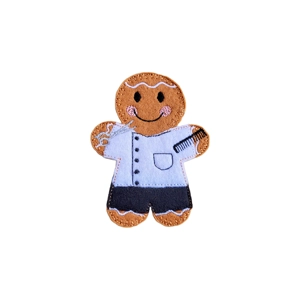 Gingerbread Barber Character made from felt, wearing a white barber’s coat and black trousers, holding a silver pair of scissors in one hand and a black comb in the other.
