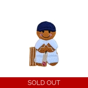Felt gingerbread cricket player character wearing a navy cap and white cricket kit, holding a bat with pads and a small red cricket ball at the feet, standing beside brown stumps.
