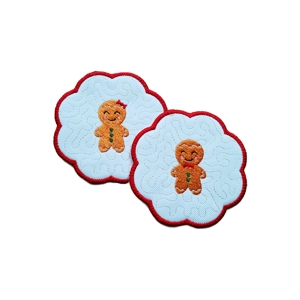 Two scalloped Christmas candle coasters featuring embroidered gingerbread characters, one gingerbread boy and one gingerbread girl, stitched on white fabric with a red border and subtle stitched pattern.