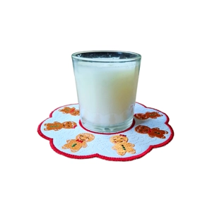 A round Christmas candle mat with embroidered gingerbread characters on a white background and a red stitched scalloped edge, shown with a glass candle placed in the centre.