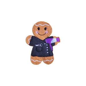 A handmade felt gingerbread hairdresser character with a stitched smile, wearing a black tunic and holding a purple and silver embroidered hairdryer.